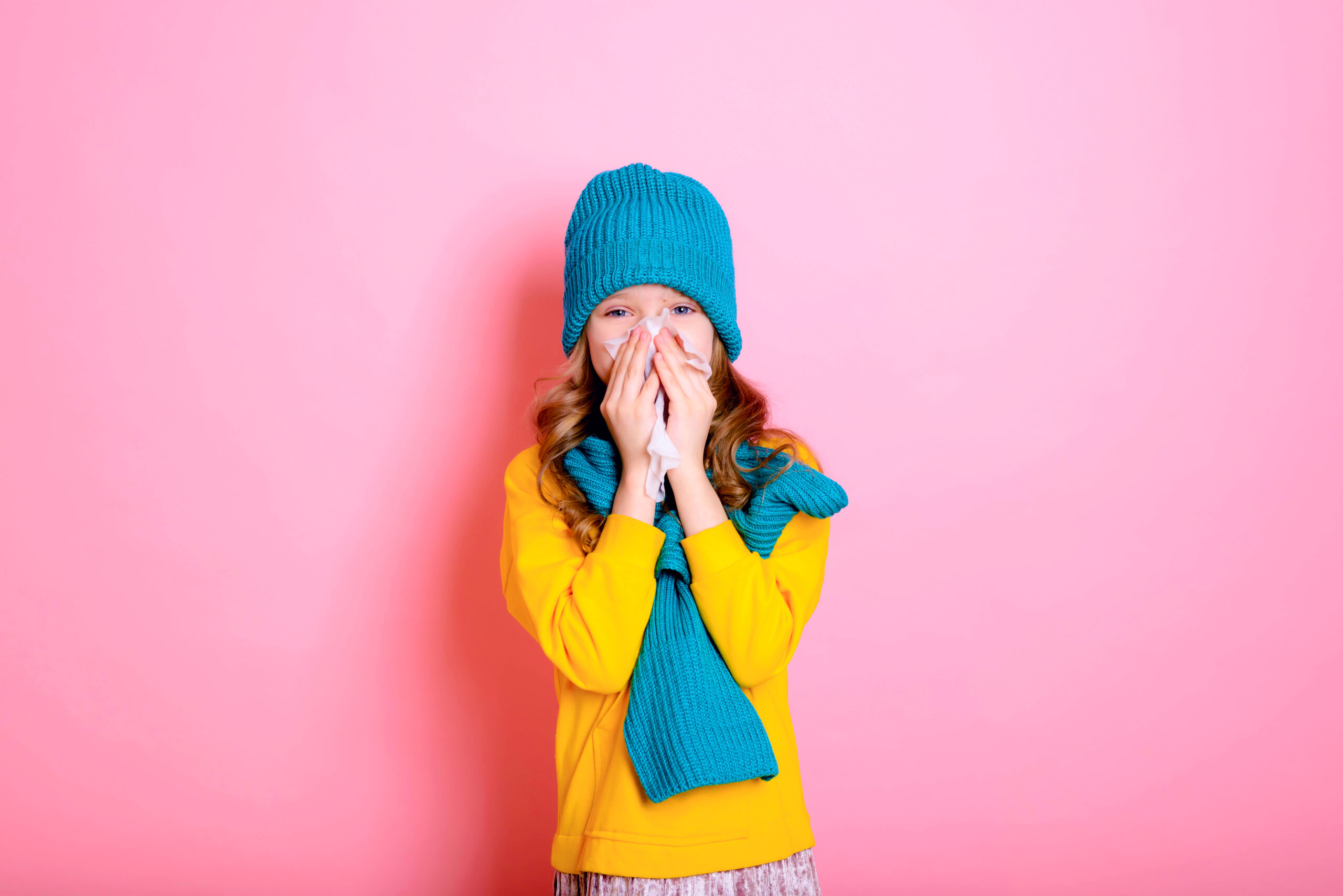 Źródło: Cold. Rhinitis. Allergy. Virus. Sneeze. Handkerchief. Blonde kid child girl is dressed in a warm sweater, scarf, and hat on a pink background. Winter and autumn seasons.